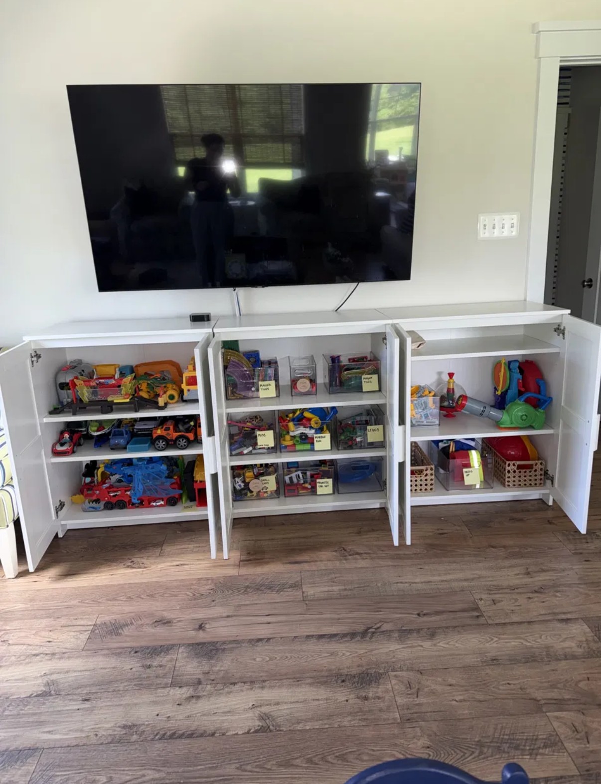 Playroom cabinet with three bays of toys sorted into labeled clear bins — Magna Tiles, Legos, cars, dress-up